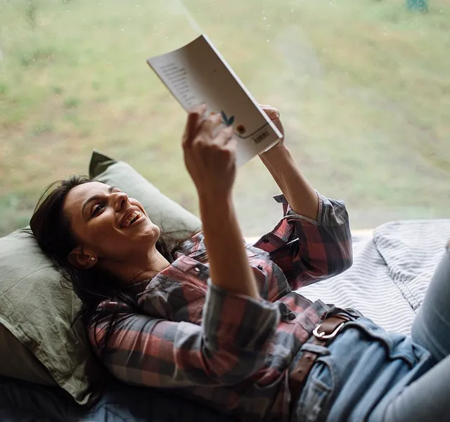 A woman reading a book while lying on a bed by a window