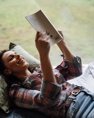 A woman reading a book while lying on a bed by a window