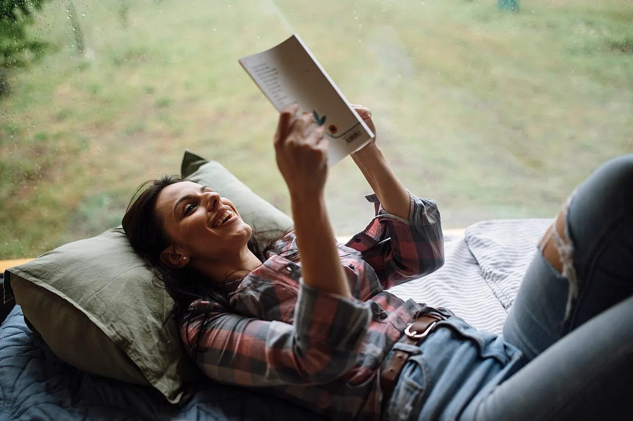 A woman reading a book while lying on a bed by a window
