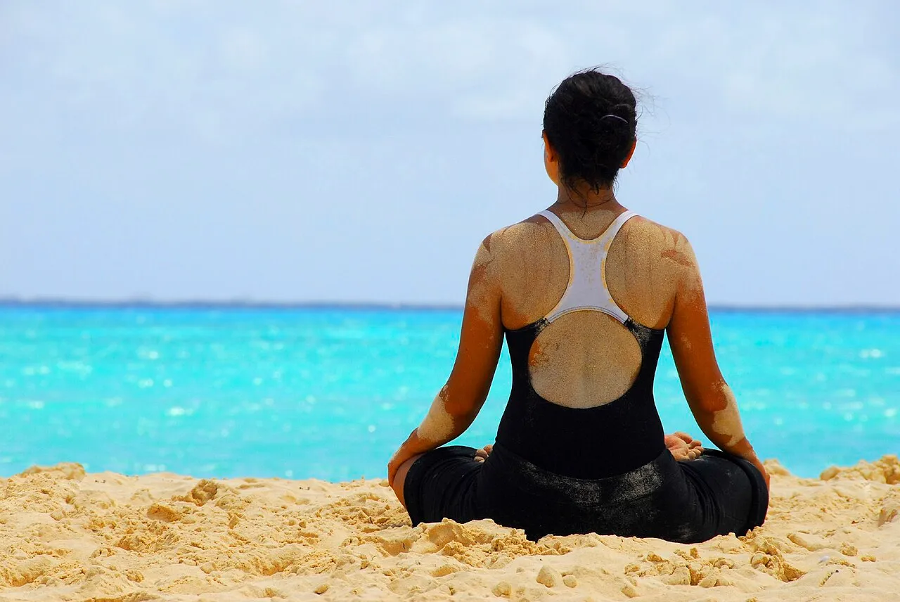 A woman seated in a meditation pose on the sand, facing turquoise water